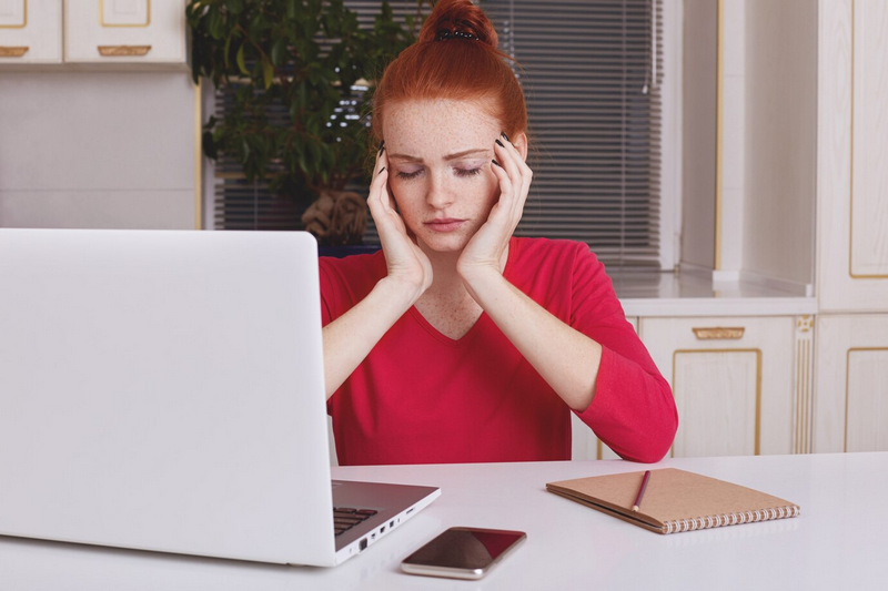 Женщина с рыжими волосами выглядит напряженной, сидя перед ноутбуком.
